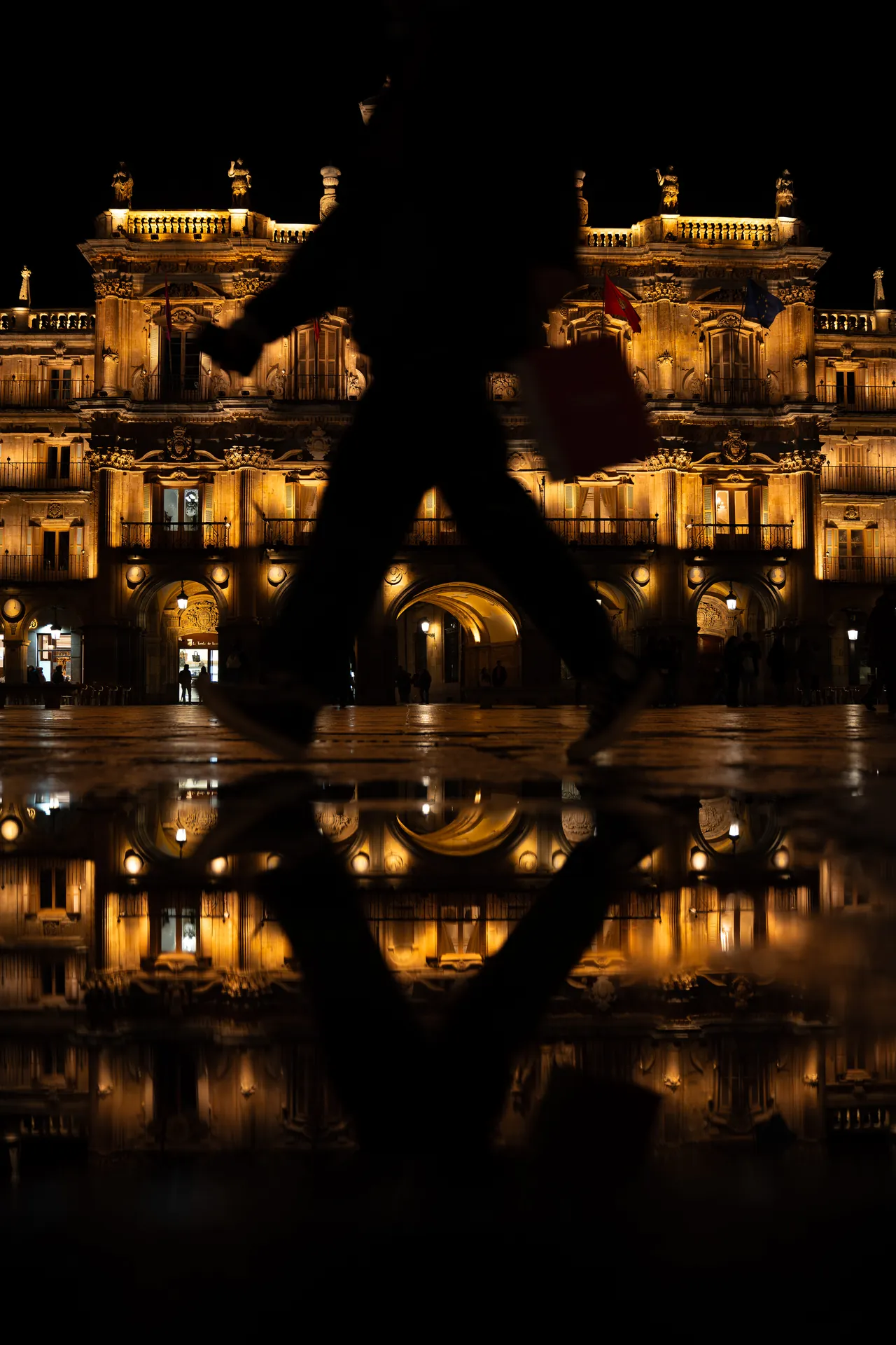 Silueta caminando ante un edificio iluminado reflejado en el suelo mojado