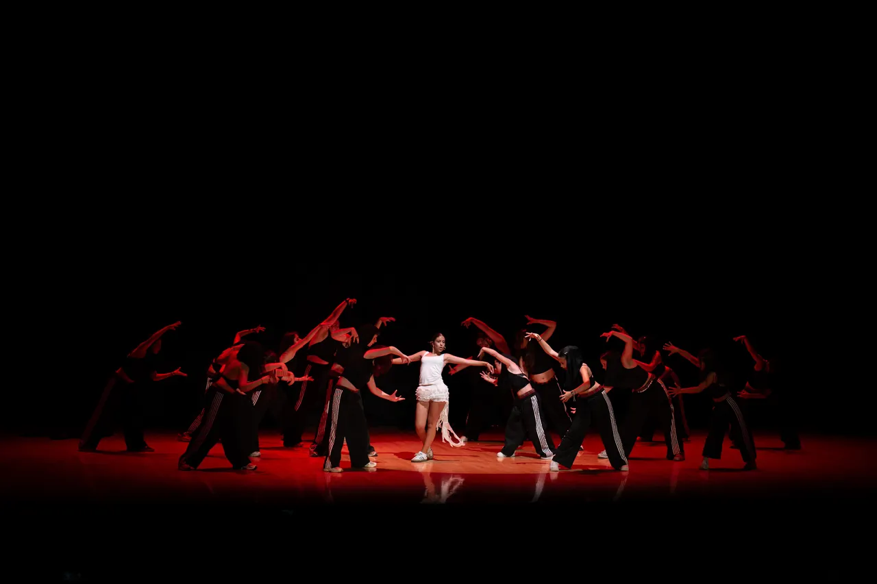 Grupo de bailarines de negro rodeando a una bailarina de blanco sobre un escenario rojo