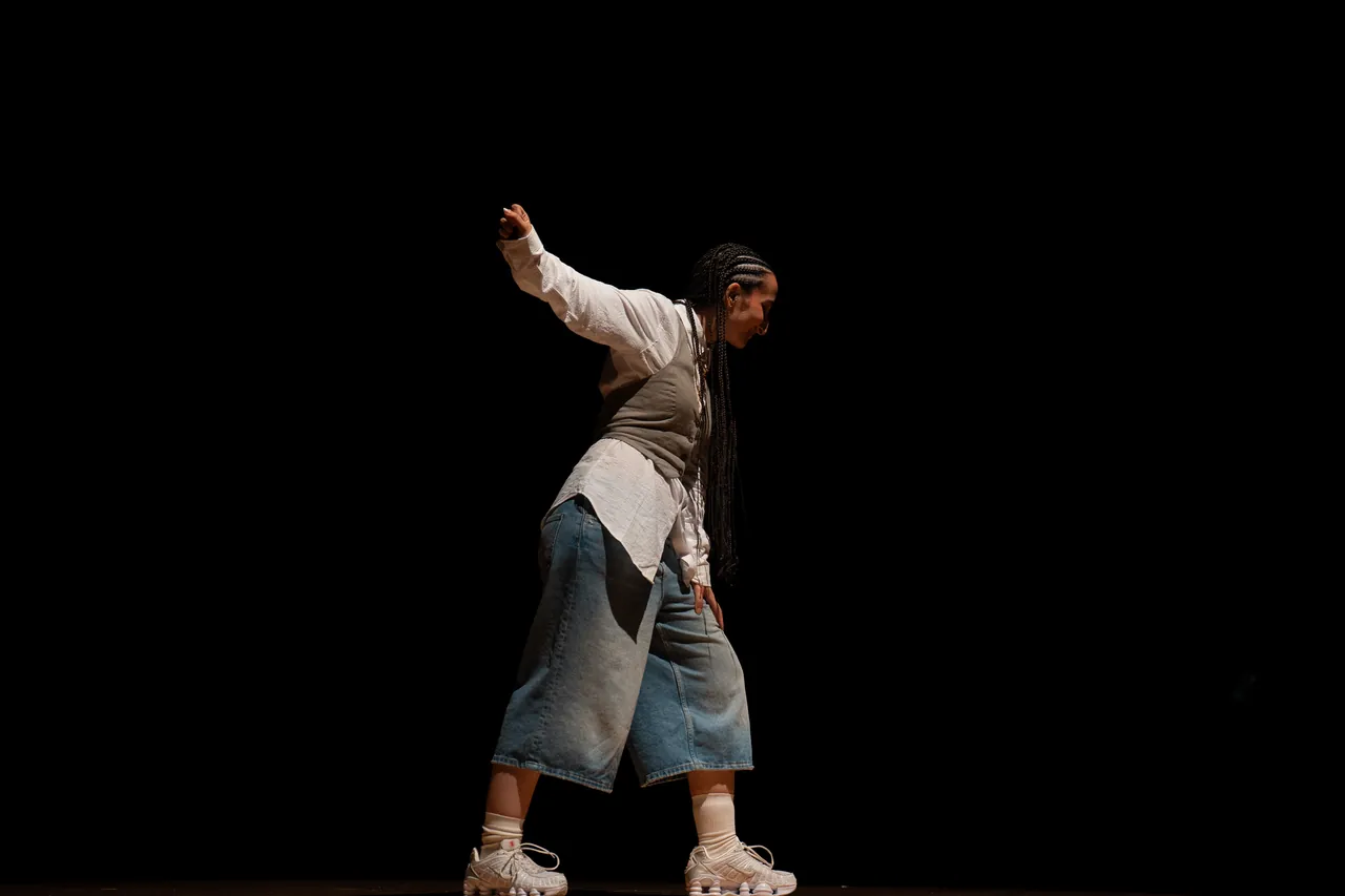 Bailarín con trenzas y vaqueros en mitad de un movimiento sobre escenario oscuro